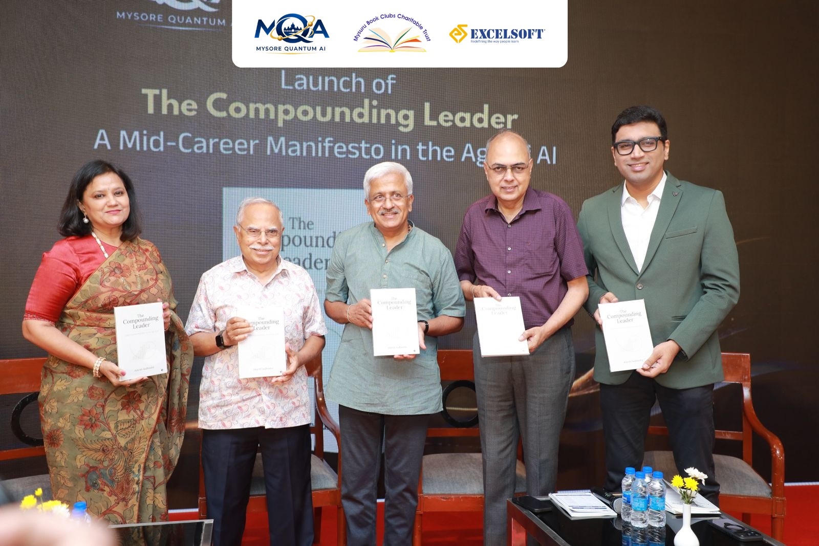 Dignitaries holding copies of The Compounding Leader on stage at the launch event in Mysuru